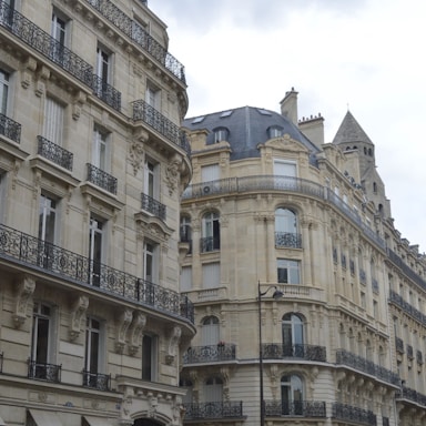 Elegant, historic buildings with intricate ironwork railings and grand facades line a street. The architecture features large windows, stone carvings, and a mixture of cream and grey tones. A tall, pointed roof structure is visible in the background.
