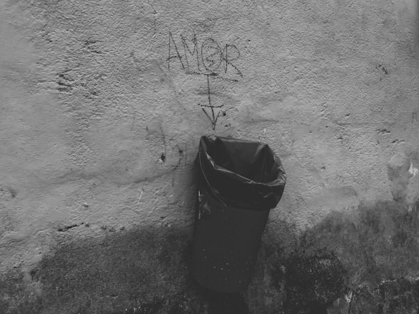 A weathered wall with graffiti text reading 'AMOR' in black, accompanied by an arrow pointing downwards. Below the graffiti, a black plastic trash bag is placed inside a metal trash can attached to the wall. The scene is monochromatic, depicting an urban and slightly gritty environment.
