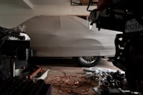 A covered car is parked in a dimly lit garage. The car is fully concealed by a light gray cover, with the shape of the vehicle and its wheels slightly visible underneath. Various automotive parts and tools are scattered on the garage floor, including a coiled extension cord and metal components.