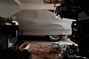 A covered car is parked in a dimly lit garage. The car is fully concealed by a light gray cover, with the shape of the vehicle and its wheels slightly visible underneath. Various automotive parts and tools are scattered on the garage floor, including a coiled extension cord and metal components.