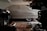 A covered car is parked in a dimly lit garage. The car is fully concealed by a light gray cover, with the shape of the vehicle and its wheels slightly visible underneath. Various automotive parts and tools are scattered on the garage floor, including a coiled extension cord and metal components.