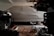 A covered car is parked in a dimly lit garage. The car is fully concealed by a light gray cover, with the shape of the vehicle and its wheels slightly visible underneath. Various automotive parts and tools are scattered on the garage floor, including a coiled extension cord and metal components.