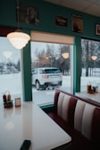 A cozy diner interior with retro-style booths and a window view of a snowy outdoor scene. The walls are decorated with vintage posters and the teal color gives a nostalgic ambiance. Outside, a white SUV is parked near some bare trees covered in snow.