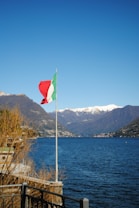 A tranquil lake with clear blue waters, surrounded by mountains topped with snow. An Italian flag waves prominently near the edge of the water, with dry shrubs and a railing in the foreground. The sky is bright and cloudless, adding to the serene atmosphere.