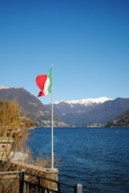 A tranquil lake with clear blue waters, surrounded by mountains topped with snow. An Italian flag waves prominently near the edge of the water, with dry shrubs and a railing in the foreground. The sky is bright and cloudless, adding to the serene atmosphere.