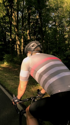 A cyclist wearing a helmet and a striped cycling jersey rides on a shaded, scenic forest road. The sun casts dappled shadows through the trees, creating a peaceful and serene atmosphere.