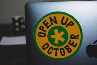 A laptop featuring a round yellow sticker with a green border and a green cross in the center. The text 'OPEN UP OCTOBER' is printed in black around the cross. The edge of an Apple logo is visible on the laptop. The background includes a blurred object.