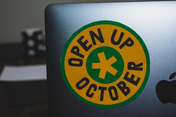 A laptop featuring a round yellow sticker with a green border and a green cross in the center. The text 'OPEN UP OCTOBER' is printed in black around the cross. The edge of an Apple logo is visible on the laptop. The background includes a blurred object.
