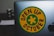 A laptop featuring a round yellow sticker with a green border and a green cross in the center. The text 'OPEN UP OCTOBER' is printed in black around the cross. The edge of an Apple logo is visible on the laptop. The background includes a blurred object.