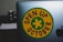 A laptop featuring a round yellow sticker with a green border and a green cross in the center. The text 'OPEN UP OCTOBER' is printed in black around the cross. The edge of an Apple logo is visible on the laptop. The background includes a blurred object.