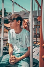 A young person with medium-length brown hair is seated outdoors, hands clasped, and looking to the side. They are wearing a white t-shirt with large printed letters and blue denim jeans. Surrounding them are metal railings, chains, and part of a rustic equipment, suggesting an industrial or construction setting. The background shows blurred trees and rooftops under a blue sky.
