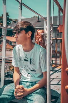 A young person with medium-length brown hair is seated outdoors, hands clasped, and looking to the side. They are wearing a white t-shirt with large printed letters and blue denim jeans. Surrounding them are metal railings, chains, and part of a rustic equipment, suggesting an industrial or construction setting. The background shows blurred trees and rooftops under a blue sky.