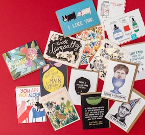 A collection of various greeting cards is displayed on a red background. The cards feature a mix of themes such as sympathy, humor, and birthday wishes. Designs include illustrated animals, flowers, and playful typography. Some cards have witty slogans, while others convey warm sentiments.