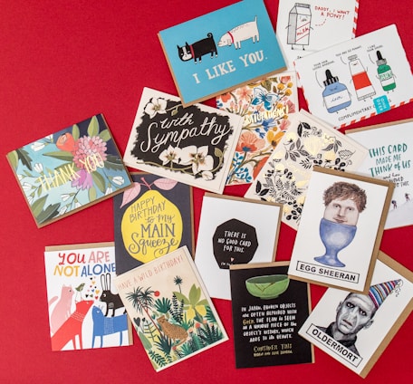 A collection of various greeting cards is displayed on a red background. The cards feature a mix of themes such as sympathy, humor, and birthday wishes. Designs include illustrated animals, flowers, and playful typography. Some cards have witty slogans, while others convey warm sentiments.