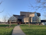 A modern architectural building with angular shapes and large glass windows is set in a grassy area. The structure features a mix of wood and metal elements. Leafless trees line the walkway leading to the entrance. People are gathered around, and a small trailer is visible to the right.