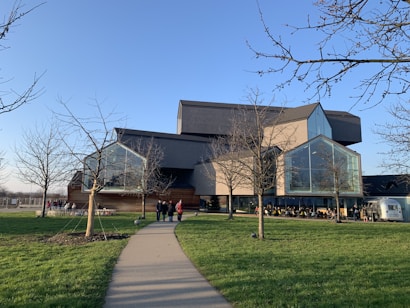 A modern architectural building with angular shapes and large glass windows is set in a grassy area. The structure features a mix of wood and metal elements. Leafless trees line the walkway leading to the entrance. People are gathered around, and a small trailer is visible to the right.
