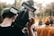 A person wearing a green beanie operates a professional video camera outdoors. The background is bokeh, with #B8C0CC figures and trees, creating a natural setting.