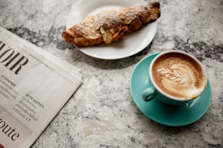 A cup of cappuccino with latte art rests on a turquoise saucer, accompanied by a croissant sprinkled with sugar on a white plate. A folded newspaper is placed nearby on a speckled granite countertop.