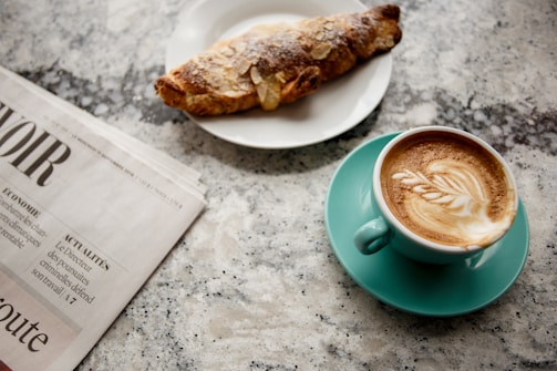 A cup of cappuccino with latte art rests on a turquoise saucer, accompanied by a croissant sprinkled with sugar on a white plate. A folded newspaper is placed nearby on a speckled granite countertop.