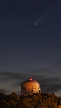 A bright comet streaking across a clear night sky over a modern industrial facility.