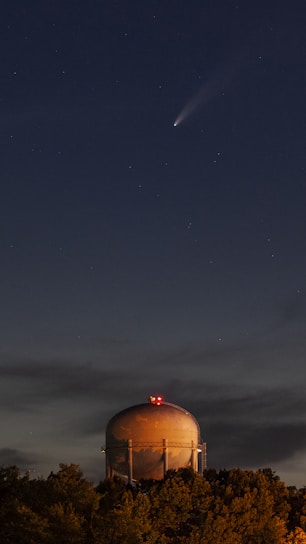 A bright comet streaking across a clear night sky over a modern industrial facility.