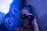 A person with long hair is wearing a sleek, fashionable outfit. They are adjusting their dark sunglasses with one hand, adorned with a black, fingerless glove. The background features a blue neon light with abstract swirling patterns, and the scene has a stylish, urban ambiance.