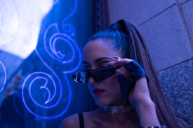 A person with long hair is wearing a sleek, fashionable outfit. They are adjusting their dark sunglasses with one hand, adorned with a black, fingerless glove. The background features a blue neon light with abstract swirling patterns, and the scene has a stylish, urban ambiance.