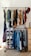 A clothes rack with two levels of hanging garments and a shoe organizer on the left. The top level contains a variety of colorful shirts and blouses, while the bottom level has jackets and jeans. The shoe organizer holds multiple pairs of shoes. A red cap is hanging on the right side of the rack, and a wooden dresser is partially visible on the right.