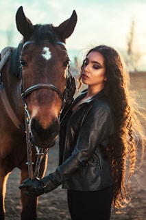 A woman with long, curly hair is standing close to a brown horse. She is wearing a black leather jacket and gloves. Her eyes are closed, and she appears calm and content. The background shows a blurred natural setting with a warm, golden light from the setting sun, creating a serene atmosphere.