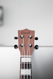 Close-up view of a guitar headstock with strings, tuning pegs, and a wooden finish. The brand name is visible on the headstock against a plain, light-colored background.