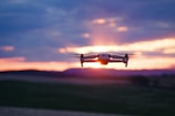 A flying drone captured against a vibrant sunset sky with hues of orange, pink, and purple. The drone is centrally positioned with blurred landscape beneath, suggesting motion and a focus on aerial capabilities.