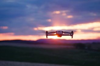 A flying drone captured against a vibrant sunset sky with hues of orange, pink, and purple. The drone is centrally positioned with blurred landscape beneath, suggesting motion and a focus on aerial capabilities.