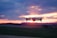 A flying drone captured against a vibrant sunset sky with hues of orange, pink, and purple. The drone is centrally positioned with blurred landscape beneath, suggesting motion and a focus on aerial capabilities.