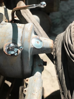A close-up of a metal mechanical component featuring rusty areas and worn surfaces. Two circular gauges or knobs are visible, along with a metallic lever extending from the top. The background includes other industrial or mechanical elements with a rough, aged texture.