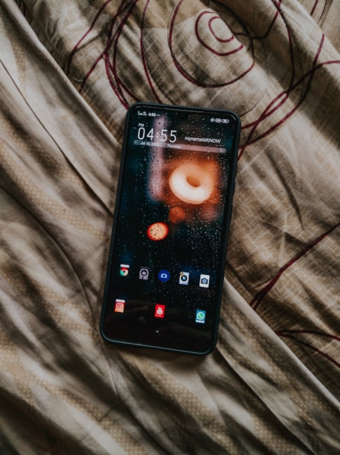 A smartphone displaying its home screen with various app icons rests on a fabric surface that has a swirling pattern. The phone screen shows droplets, suggesting a rain effect or theme.