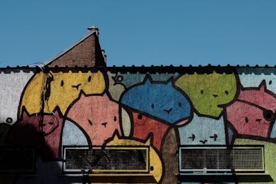 Colorful mural featuring cartoon-like cats in various hues, covering a wall with metal grates on the lower section. The cats have simplistic, smiling faces and are rendered in bold outlines and bright colors.