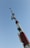 A tall communication tower with multiple antennas and dishes extends upwards, set against a clear blue sky. The structure is painted in red and white with visible cables and equipment attached.