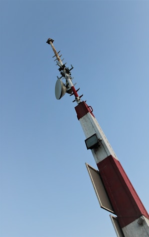 A tall communication tower with multiple antennas and dishes extends upwards, set against a clear blue sky. The structure is painted in red and white with visible cables and equipment attached.