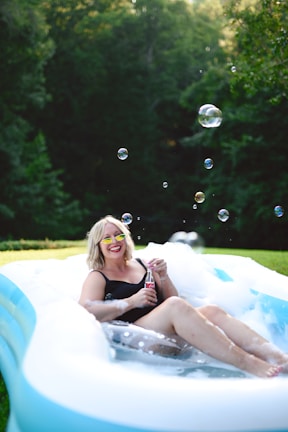 A person wearing a black swimsuit and sunglasses is sitting and smiling in an inflatable pool filled with bubbles. The individual is holding a bottle of soda and several soap bubbles are floating in the air. The background features lush green trees and grass, creating a serene outdoor setting.