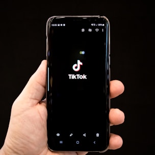 A hand holds a smartphone displaying the TikTok logo on the screen. The phone's screen is predominantly black, highlighting the logo in the center. The background is also dark, creating a focus on the phone and the hand.