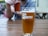 A glass of amber beer with a frothy head is placed on a wooden table. The glass has the logo of Madtree Brewing Company. In the background, a person holds another similar glass. The setting appears to be a casual outdoor environment, possibly a beer garden or patio.