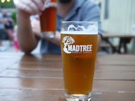 A glass of amber beer with a frothy head is placed on a wooden table. The glass has the logo of Madtree Brewing Company. In the background, a person holds another similar glass. The setting appears to be a casual outdoor environment, possibly a beer garden or patio.