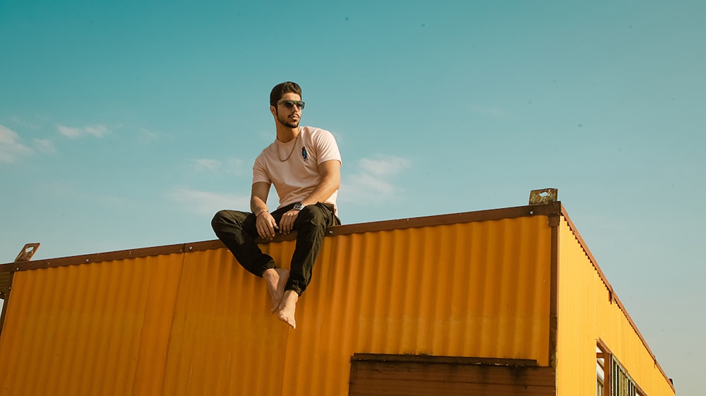 man in white crew neck t-shirt and black pants sitting on yellow wall during daytime