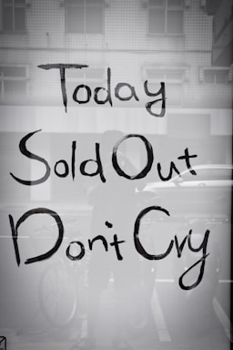 A shop window displays text reading 'Today Sold Out Don't Cry' in a casual, hand-painted style. The reflection shows part of a bicycle and some indistinct elements in an urban setting.