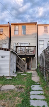 A three-story townhouse with beige siding, featuring a small porch on the second floor and a set of stairs leading down to the ground level. The house is part of a row of similar attached homes, with a concrete and grass pathway leading to the entrance. The sky is cloudy with patches of blue visible.