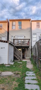 A three-story townhouse with beige siding, featuring a small porch on the second floor and a set of stairs leading down to the ground level. The house is part of a row of similar attached homes, with a concrete and grass pathway leading to the entrance. The sky is cloudy with patches of blue visible.
