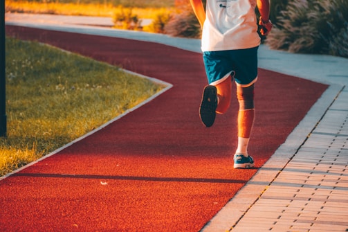A person in athletic attire is jogging on a red running track that curves through a park. The runner is wearing blue shorts, a white shirt, and blue running shoes. Sunlight casts a warm glow over the scene, and the path is surrounded by grassy areas.
