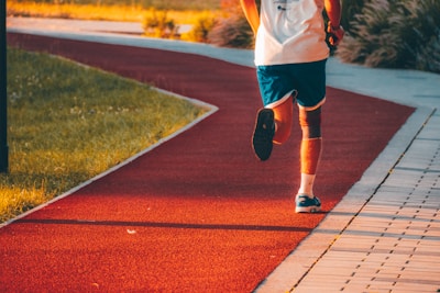 A person in athletic attire is jogging on a red running track that curves through a park. The runner is wearing blue shorts, a white shirt, and blue running shoes. Sunlight casts a warm glow over the scene, and the path is surrounded by grassy areas.