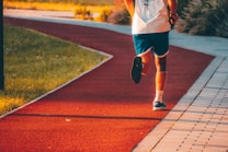 A person in athletic attire is jogging on a red running track that curves through a park. The runner is wearing blue shorts, a white shirt, and blue running shoes. Sunlight casts a warm glow over the scene, and the path is surrounded by grassy areas.