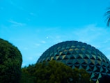 A geodesic dome structure is partially visible surrounded by trees under a clear blue sky. The geometric patterns on the dome are reflective, creating a mix of dark and light shades. There is a small bright object in the sky, possibly the moon.